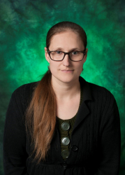 Picture of Staff Senate Katie Hebert. Katie has dark glasses, brown colored eyes and a long brown ponytail. Katie is against a green background and is wearing a black blazer with large circular buttons in the center