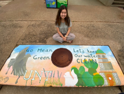 A student artists sits next to her storm drain mural depicting UNT iconic imagery and a sunset