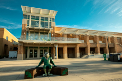 Scrappy the Eagle mascot sitting in front of the University Union building on a brown bench with green eagles