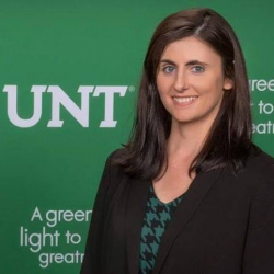 Picture of Claudia Taylor. Claudia has shoulder length brown hair and green eyes. Claudia is wearing a green patterned shirt with a black blazer and is standing against a green UNT background.