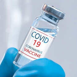 Picture of COVID-19 vaccine in clear vial with label being held by a medical professional wearing a blue glove