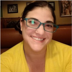 Picture of Staff Success Stories Winner Diana Bergeman. Diana is in a restaurant booth with framed pictures in the background. Diana has brown hair that is pulled back, blue glasses and a yellow top