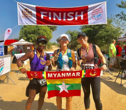 Picture of Janis Miller (center). Janis completed a marathon in Myanmar and is posing with two other runners. The runners are standing in front of the "Finish" sign and holding a Myanmar flag