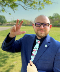 Picture of Vince Fisher. Vince has a bald head, black glasses and his holding his left hand in a talon position. Vince is wearing a blue suit with a North Texas pin, black shirt and pastel blue tie with a floral pattern