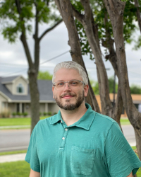 Picture of Staff Success Stories Winner William Schuelke. William is outside in a yard with a house and tree in the background. William has gray hair with a black beard and is wearing a green polo